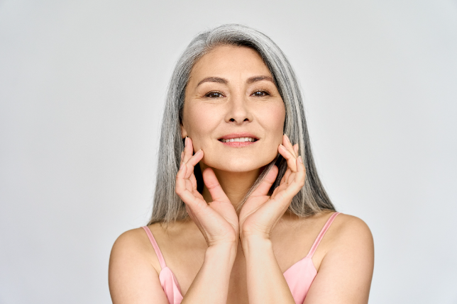 A confident mature woman with grey hair smiles while gently touching her cheeks, showcasing the results of skin tightening therapy.